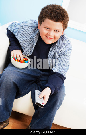 Il sovrappeso boy (13-15) di mangiare cibo spazzatura, tenendo il controllo remoto Foto Stock