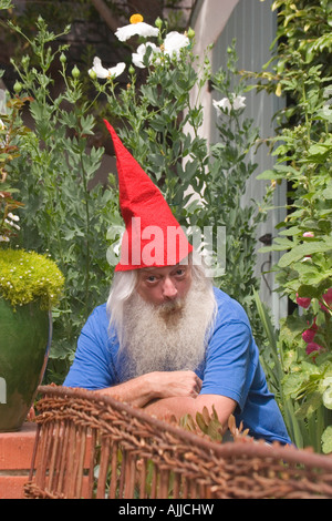 Un uomo vestito come un barbuto gnome indossando una punta di Red Hat, si accovaccia in un giardino con una premurosa espressione sul suo volto. Foto Stock