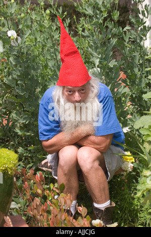 Un uomo vestito come un barbuto gnome indossando una punta di Red Hat, si accovaccia in un giardino con una premurosa espressione sul suo volto. Foto Stock