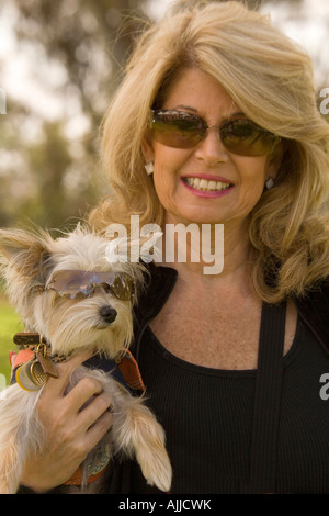 Una donna di mezza età sorrisi e mantiene il suo amato piccolo cane. Entrambi sono indossando occhiali da sole. Foto Stock