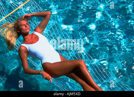 Donna bionda in costume da bagno bianco giacente su amaca su acque blu cristallo di una piscina Foto Stock