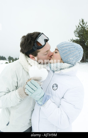 Coppia giovane kissing, tenendo il cuore in neve insieme a occhi chiusi, ritratto Foto Stock