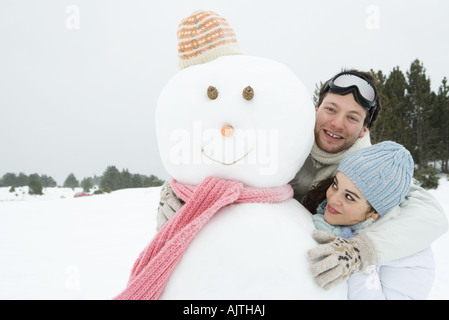 Due giovani amici in piedi con pupazzo di neve, entrambi sorridenti, uno che guarda in telecamera Foto Stock