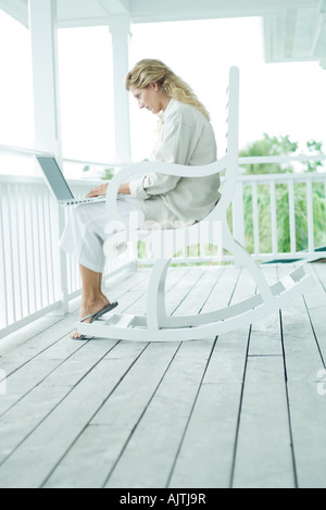 Donna seduta in sedia a dondolo sulla veranda, utilizzando il computer portatile a piena lunghezza Foto Stock