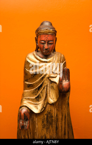 In legno statua di Buddha gesticolando con una mano e una mano verso il basso contro la parete arancione Foto Stock