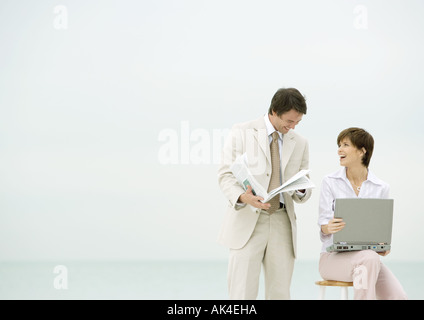 Due persone di affari sulla spiaggia, uno utilizzando laptop, l'altra lettura giornale Foto Stock