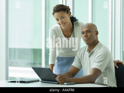 Imprenditore seduto alla scrivania mentre la donna si appoggia sulla spalla, sorridente in telecamera Foto Stock