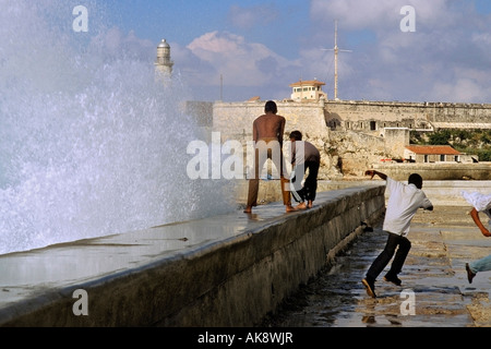 Ragazzi giocare in onde che si infrangono lungo il Malecón in Havana Cuba con la fortezza El Morro in background Foto Stock