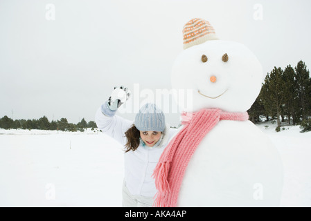 Giovane donna in piedi accanto al pupazzo di neve, pronto a lanciare palle di neve in telecamera Foto Stock