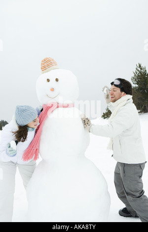 Coppia giovane peeking intorno pupazzo di neve a ogni altra azienda fino snowballs Foto Stock