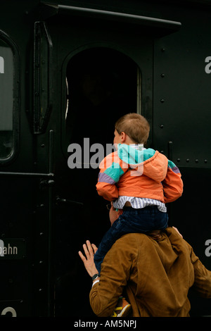 Uomo con un bambino piccolo sulle sue spalle guarda nella cabina di uno storico treno a vapore Foto Stock