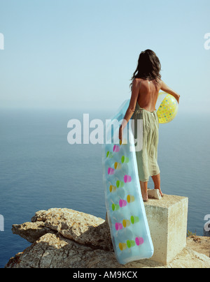 Donna in piedi sulla roccia che guarda sul mare tenendo una palla spiaggia e a lilo. Foto Stock