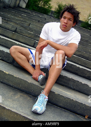 Ritratto ambientale di un esotico uomo seduto sui gradini indossa pantaloncini bianchi camicia bianca e scarpe da tennis blu sneakers Foto Stock