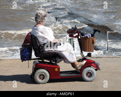 Lungomare lungomare, donna matura che guida su scooter motorizzato per disabili, che osserva la marea dura e alta Walton sul Naze Essex Inghilterra, Regno Unito Foto Stock