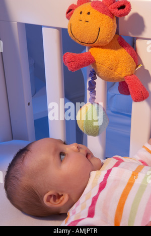 Un bambino giace sulla sua schiena in una culla e guarda ad un mobile in un vivaio di notte Foto Stock