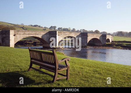 Banco vuoto con vista fiume Wharfe e pietra ponte arcuato in villaggio in Yorkshire Dales National Park. Wharfedale Burnsall Yorkshire England Regno Unito Foto Stock