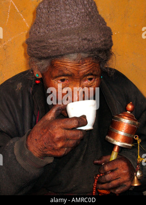 Donna anziana annerito con fuliggine sips tè durante la filatura ruota di preghiera Lhatse Shekor Tibet Foto Stock