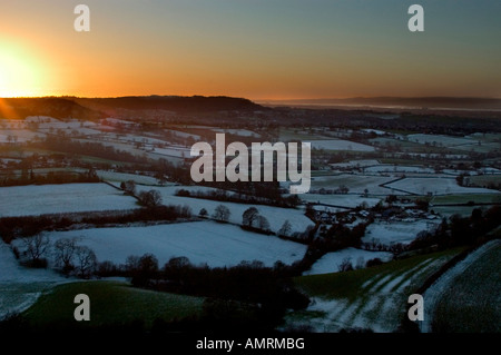 Winter scene with snow covered fields, woods and hedges with sun setting behind  distant hill across Vale of Gloucester Foto Stock