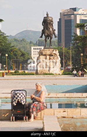 La statua del xv secolo un guerriero e un eroe nazionale Skanderbeg Skanderburg su un enorme base di pietra. Una donna seduta sul davanzale di una fontana con un bambino la PRAM. Il Tirana Piazza Centrale, Skanderburg Skanderbeg Square. Capitale Tirana. L'Albania, Balcani, Europa. Foto Stock