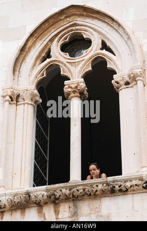 Una giovane donna che guarda fuori dal Palazzo del Rettore Knezev Dvor attraverso un arcuato finestra gotica, guardando annoiato contro lo sfondo nero sul Pred Dvorom street Dubrovnik, città vecchia. Costa della Dalmazia, Croazia, Europa. Foto Stock