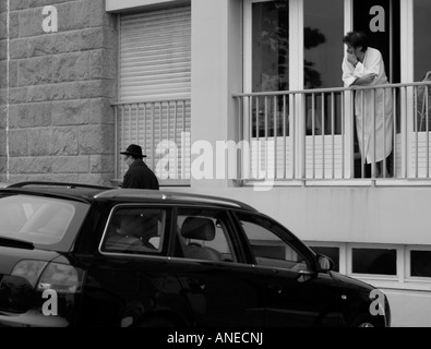 La donna in asciugamano bianco accappatoio appoggiata al suo ringhiera di balcone a guardare un uomo che indossa un cappello nero a piedi dalla provinciale in Francia Foto Stock