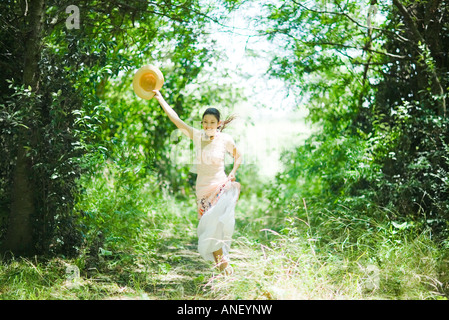 Giovane donna in sundress che corre lungo il percorso boschivo, tenendo alto cappello per il sole Foto Stock