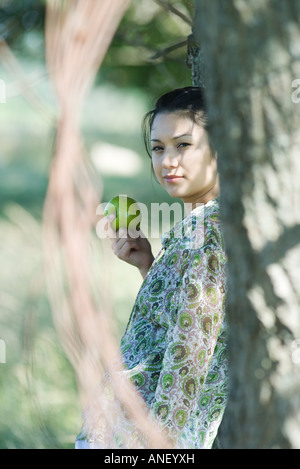 Giovane donna appoggiata contro l'albero, in possesso di Apple Foto Stock