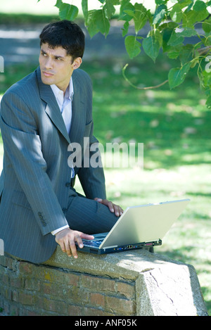 Imprenditore seduto sul muro basso nel parco, utilizzando laptop, guardando lontano Foto Stock