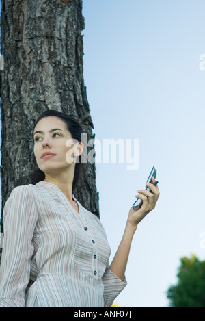 Donna che mantiene il telefono cellulare, appoggiata ad albero, a basso angolo di visione Foto Stock