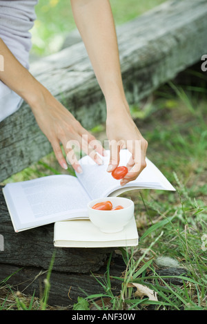 Donna libro di lettura e di mangiare i pomodori ciliegia, ritagliato Foto Stock