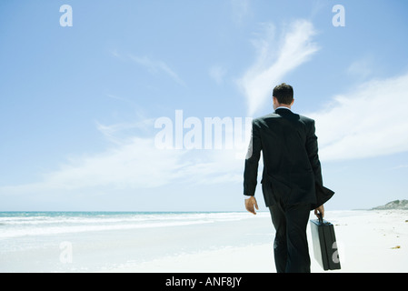 Imprenditore camminando di fronte spiaggia, portando nella valigetta, vista posteriore Foto Stock