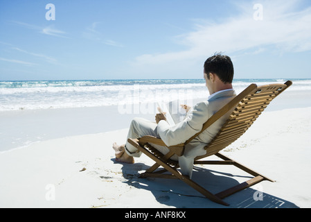 Imprenditore seduto nella sedia a sdraio sulla spiaggia, libro di lettura Foto Stock