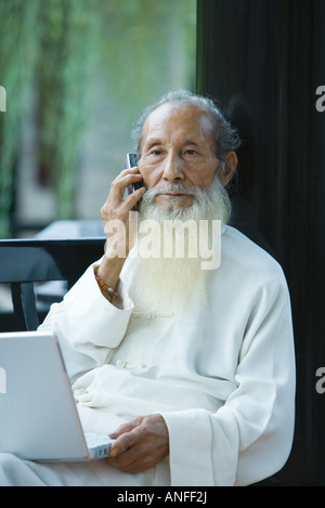 Uomo anziano che indossa il cinese tradizionale abbigliamento, utilizzando computer portatile e un telefono cellulare Foto Stock