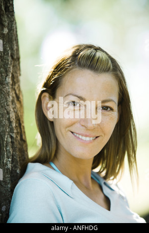 Donna appoggiata ad albero, sorridente in telecamera, ritratto Foto Stock