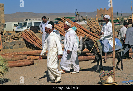 Gli uomini locali in bianco tradizionale djellabahs passeggiate a piedi e a cavallo di asino passato un timberyard al deserto città mercato di Zagora, sud del Marocco Foto Stock