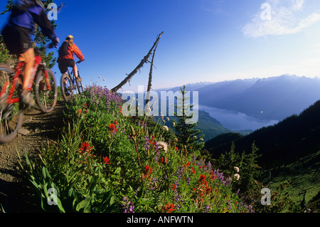 Due woman mountain bikers sul picco di Idaho con Valhallas dietro, Slocan Valley, Kootenays, British Columbia, Canada. Foto Stock