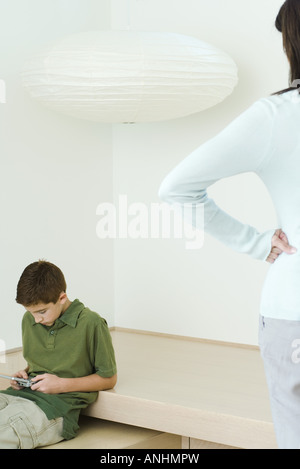 Ragazzo la riproduzione di video gioco, madre a guardare le mani sui fianchi Foto Stock