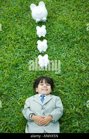 Little Boy in piena suit giacente su erba, linea di piggy banche sopra la testa Foto Stock