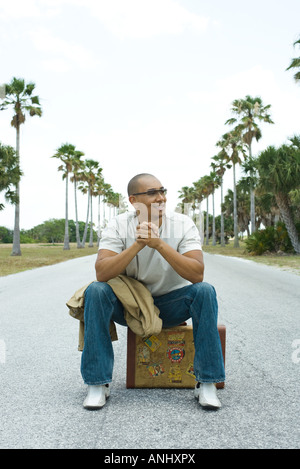 Uomo seduto sulla valigia nel mezzo della strada, sorridente, guardando lontano Foto Stock
