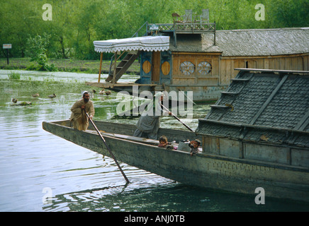 Una famiglia che punisce la loro casa galleggiante tradizionale attraverso il lago dal con una pittoresca casa galleggiante in affitto sullo sfondo. Srinagar, Kashmir. India Foto Stock