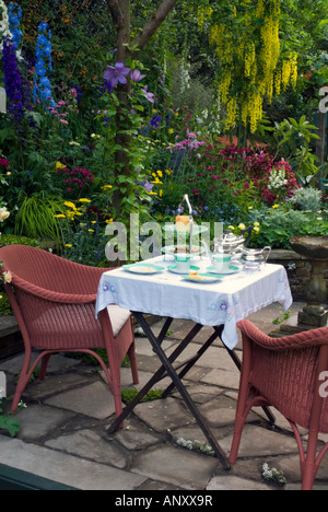 Lussureggiante giardino set per mangiare un tè o per la colazione con sedie e tavolo da pranzo sul patio, riempito con fiori colorati e piante Foto Stock