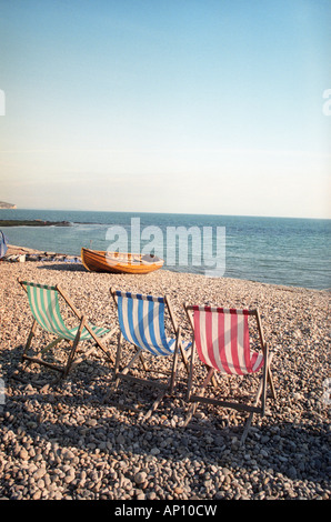 Tre vivacemente colorati di sedie a sdraio e la barca di legno su una spiaggia ghiaiosa Foto Stock