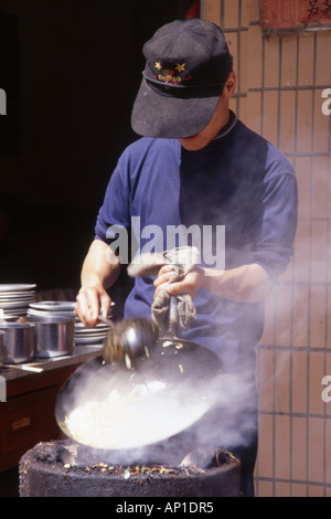 Uomo locale indossando cappello da baseball per la cottura di alimenti in un wok fumante,Chengdu,provincia di Sichuan, in Cina. Foto Stock
