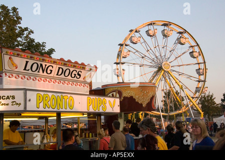 Testa e spalle colpo di Fair patroni fresatura circa & in attesa in un hot dog stand per il cibo. Una ruota panoramica Ferris serve come sfondo. Foto Stock