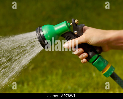 Mano femminile che tiene in mano una pistola a spruzzo con tubo flessibile e innaffia un prato in una calda giornata di sole nel Regno Unito Foto Stock