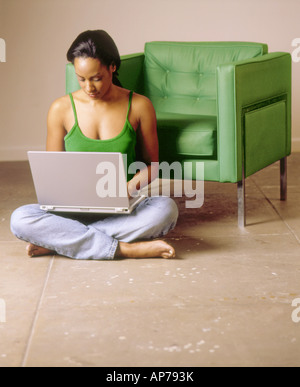 Americano africano nero donna si siede sul pavimento con verde sedia moderna in camera con il computer portatile di lavoro o di shopping online Foto Stock