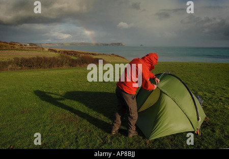 Un uomo pitching una tenda in un campeggio sulla Costa in Cornwall, Regno Unito, nel vento cattivo tempo piovoso, indossando un impermeabile rosso Foto Stock
