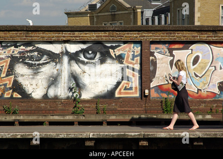 Una giovane donna solitaria che cammina da sola sulla piattaforma della stazione ferroviaria di South London, con opere d'arte graffiti dipinte su un muro nella soleggiata Londra meridionale, Inghilterra, Regno Unito Foto Stock