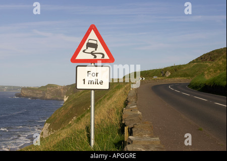 Triangolare foderato rosso strada sdrucciolevole per un miglio avvertenza cartello stradale sulla A2 coast road vicino a piegare con Dunluce Castle nel retro Foto Stock