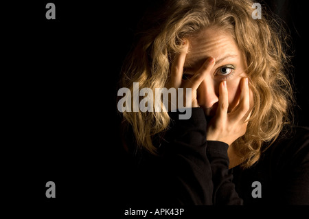 Giovane donna bionda che tiene il capo nelle sue mani in studio aginst sfondo nero fa capolino tra le dita Foto Stock